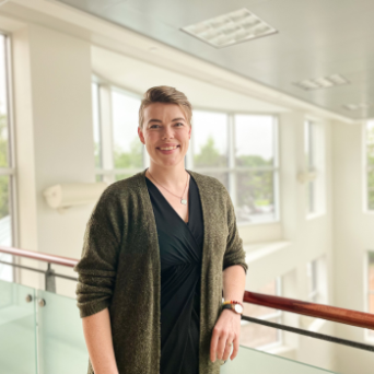 Emma has light skin and short brown hair. She stands with her arm resting on a railing inside a sunny room. She wears a black shirt and green sweater.